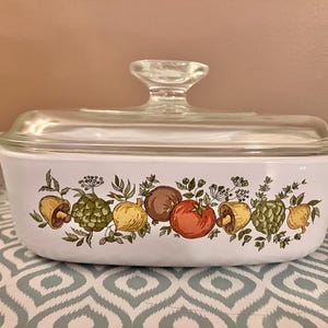 Puede incluir: Fuente rectangular blanca con tapa de cristal transparente. La fuente está decorada con un colorido estampado de verduras, como tomates, champiñones y alcachofas. La tapa tiene un asa de cristal transparente.