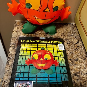 May include: An orange inflatable pumpkin with a smiling face and green feet, packaged in a plastic bag labelled 14" 35.6cm INFLATABLE PUMPKIN. The packaging features a grid background, a small photo of people in costumes, and the text Home Decoration Made Of Soft, Safe Vinyl.