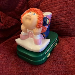 Puede incluir: Una figura de Cabbage Patch Kids de una niña con cabello rojo que lleva auriculares y está acostada sobre una almohada. La figura está sobre una base verde con las palabras "Cabbage Patch Kids" impresas en ella.