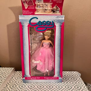 Peut inclure: Une poupée rose et blanche avec des cheveux blonds et une robe rose. La poupée est emballée dans une boîte rose avec les mots "Cassy Getting Ready Doll" sur le devant.