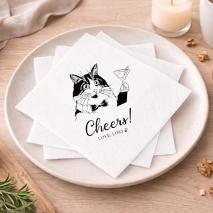 May include: White square paper napkins with a black and white illustration of a cat holding a martini glass. The napkins have the word "Cheers!" and the text "LOVE, LOKI" printed on them. The napkins are stacked on a white plate.