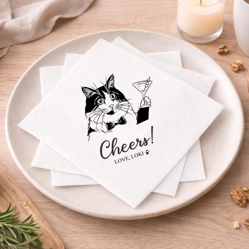 May include: White square paper napkins with a black and white illustration of a cat holding a martini glass. The napkins have the word "Cheers!" and the text "LOVE, LOKI" printed on them. The napkins are stacked on a white plate.