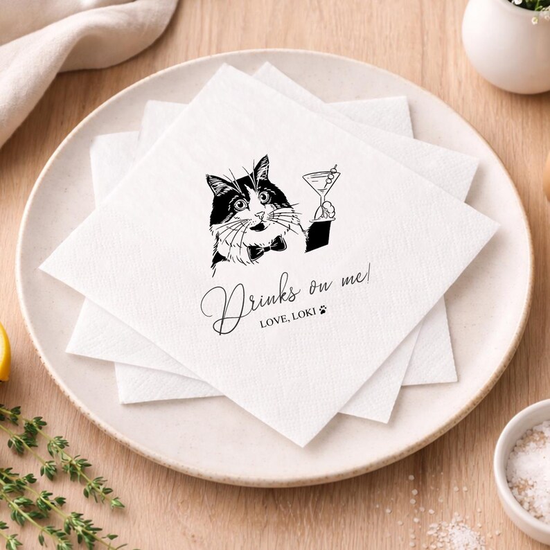May include: White square paper napkins with a black and white illustration of a cat holding a martini glass. The text "Drinks on me!" and "LOVE, LOKI" are printed on the napkins. The napkins are stacked on a white plate.