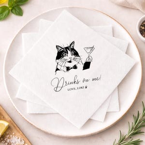 May include: White cocktail napkins with a black and white illustration of a cat holding a martini glass. The text "Drinks on me! Love, Loki" is printed below the cat. The napkins are stacked on a white plate.