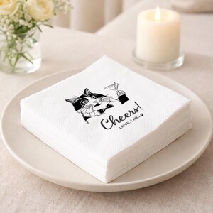 May include: A stack of white paper napkins with a black and white cat illustration holding a martini glass. The napkins have the word "Cheers!" and "LOVE, LOKI" printed on them. The napkins are on a white plate.