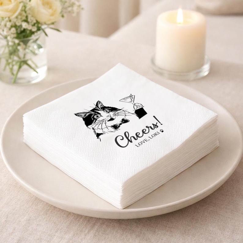 May include: A stack of white paper napkins with a black and white cat illustration holding a martini glass. The napkins have the word "Cheers!" and "LOVE, LOKI" printed on them. The napkins are on a white plate.