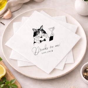 May include: White square paper napkins stacked on a plate. Each napkin features a black and white illustration of a cat holding a martini glass, with the text "Drinks on me!" and "LOVE, LOKI" in a script font.