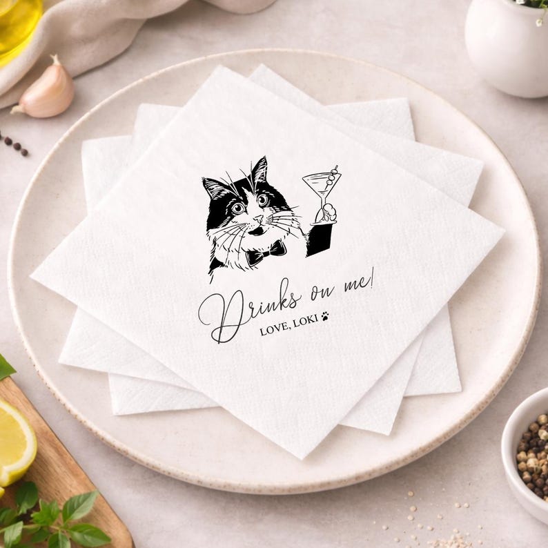 May include: White square paper napkins stacked on a plate. Each napkin features a black and white illustration of a cat holding a martini glass, with the text "Drinks on me!" and "LOVE, LOKI" in a script font.