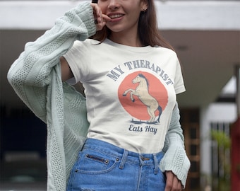 May include: A white t-shirt with a graphic of a horse and the text "My Therapist Eats Hay". The woman is wearing blue denim shorts and a light green cardigan.