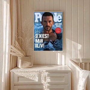 May include: A framed magazine cover featuring Jonathan Bailey, with the headline "Sexiest Man Alive." The People magazine cover is displayed on a white wall, with a neutral-toned dresser and decorative plant in the foreground.