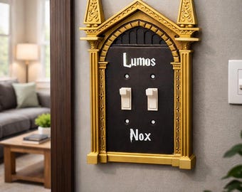 May include: A decorative light switch cover with a gold-colored frame and a black center. The words "Lumos" and "Nox" are written in white script above and below two light switches. The frame has ornate architectural details.