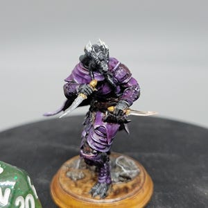 May include: A hand-painted miniature figurine of a purple and black dragon-like creature holding two daggers. The creature is wearing armour and a purple cloak. The figurine is standing on a brown base.