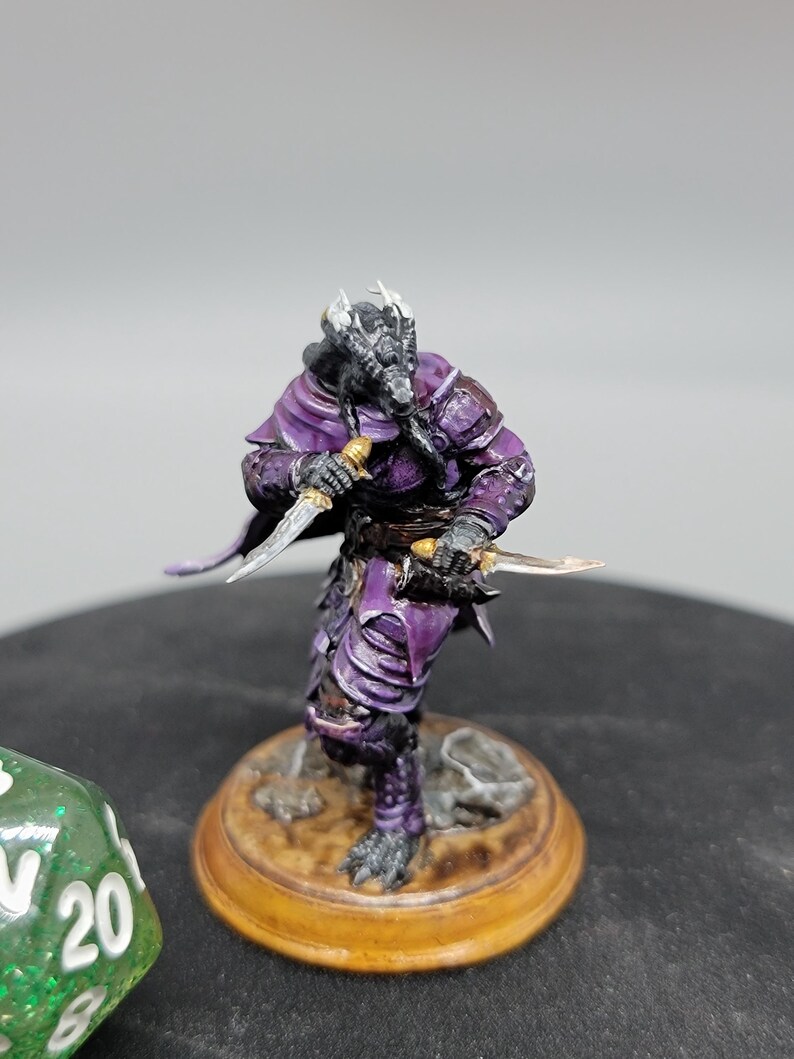 May include: A hand-painted miniature figurine of a purple and black dragon-like creature holding two daggers. The creature is wearing armour and a purple cloak. The figurine is standing on a brown base.
