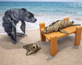 May include: Three toy seals on a sandy beach with the ocean in the background. One seal stands upright, another rests on a small wooden dock, and a third lies on the sand. The seals are gray and tan.