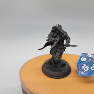 May include: A black and grey miniature figurine of a humanoid creature with a reptilian head, holding a dagger in each hand. The figurine is standing on a black base and is posed in a fighting stance.