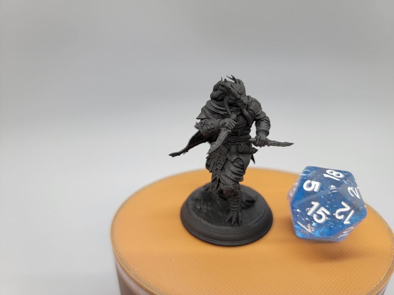 May include: A black and grey miniature figurine of a humanoid creature with a reptilian head, holding a dagger in each hand. The figurine is standing on a black base and is posed in a fighting stance.