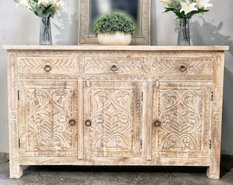 May include: A white wooden cabinet with three doors and two drawers. The cabinet has intricate carvings of flowers and leaves. There are two vases with white flowers and a potted plant on top of the cabinet. A silver framed mirror hangs on the wall above the cabinet.