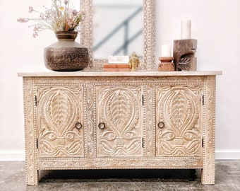 May include: A white wooden cabinet with three carved doors and a carved top. The cabinet is sitting on a wooden floor. There is a mirror and a vase with flowers on top of the cabinet.