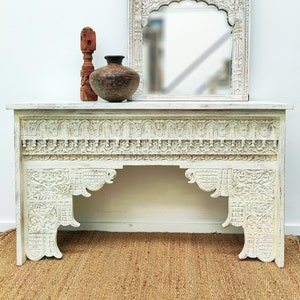 May include: White carved wooden console table with intricate details. The table has a rectangular top and two arched legs. A small wooden sculpture and a brown clay pot are on top of the table.