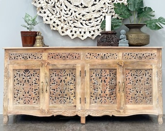 May include: A carved wooden cabinet with four doors and three drawers. The cabinet is made of light wood and has a distressed finish. The doors and drawers have intricate carvings of flowers and leaves. The cabinet is sitting on a concrete floor in front of a white wall. A large round wooden wall decor with intricate carvings is hanging on the wall above the cabinet.