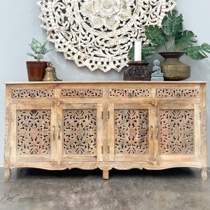 May include: A carved wooden cabinet with four doors and three drawers. The cabinet is made of light wood and has a distressed finish. The doors and drawers have intricate carvings of flowers and leaves. The cabinet is sitting on a concrete floor in front of a white wall. A large round wooden wall decor with intricate carvings is hanging on the wall above the cabinet.