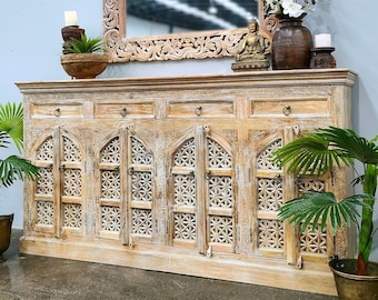 May include: A large, light-colored wooden cabinet with intricate carved details and arched doors. Above, a matching carved mirror reflects the room. The cabinet is decorated with plants, candles, and a small Buddha statue, creating a serene atmosphere.