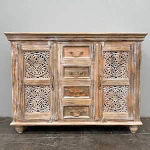May include: A light-coloured wooden cabinet with intricate carved floral designs on the doors and sides. The cabinet has five drawers and brass-coloured hardware. The cabinet is approximately 152 cm wide.