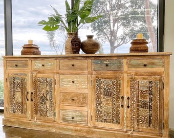 May include: A large, rustic wooden cabinet with carved doors and drawers. The cabinet is painted in a light wood stain and has a distressed finish. It features multiple drawers and doors with intricate carvings. The cabinet is sitting on a concrete floor in front of a large window.