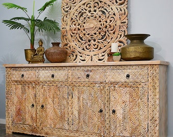 May include: A white wooden cabinet with intricate carvings and four doors. The cabinet has three drawers above the doors. The cabinet is decorated with a carved wooden wall hanging with a floral design.