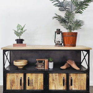 May include: An industrial-style cabinet in black and wood tones. It features four doors with wooden panels and a light wood top. The cabinet is decorated with plants, a lantern, and a wooden bowl. Ideal for storage and display, it measures approximately 150cm in length.