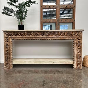 May include: A whitewashed wood console table with intricate carvings. The table has a single shelf and is made of reclaimed wood.