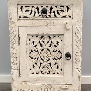 May include: White wooden bedside table with a carved floral design, a single drawer, and a cabinet door with a metal ring handle. The bedside table has a distressed white finish and sits on four carved legs.