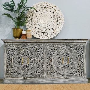 May include: A grey carved wooden cabinet with two doors and intricate floral designs. The cabinet has a distressed white paint finish and gold metal handles. A white carved wooden round wall decor is above the cabinet.