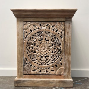 May include: A whitewashed wooden cabinet with a carved floral design on the door. The cabinet has a simple, elegant design and would be a great addition to any home.