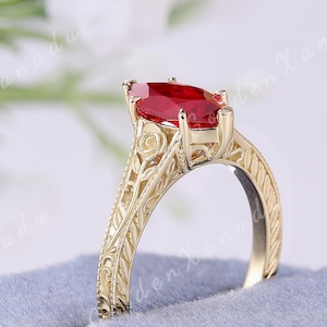 May include: A gold ring with a marquise-cut red gemstone. The ring has an ornate, engraved design on the band. The ring is displayed on a blue velvet ring box. The text "GOLDENXANADU" is at the top of the image.
