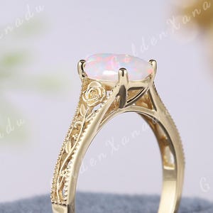 May include: A gold-colored ring with an oval, opalescent gemstone. The ring features intricate floral and leaf detailing on the band and around the gemstone setting. The ring is displayed on a blue velvet surface.