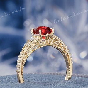 May include: A gold ring with a red gemstone and intricate filigree detailing. The ring is set on a blue velvet box.