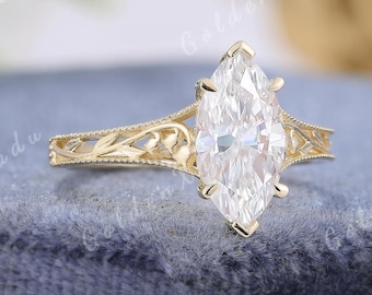 May include: A marquise-cut diamond engagement ring with a gold band. The band features an intricate filigree design with floral and leaf patterns. The ring is set on a blue velvet surface.