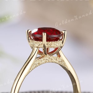 May include: A gold ring with a red gemstone set in a delicate, ornate setting. The ring is displayed on a gray velvet box.