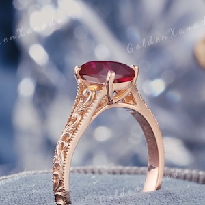 May include: A rose gold ring featuring a deep red oval-cut gemstone held by four prongs. The band displays intricate scrollwork engravings along the sides. The text GOLDENXANADU is visible at the top, with the brand name repeated in the background.