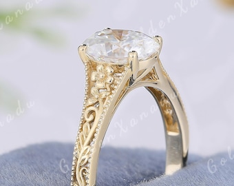 Puede incluir: Un anillo de compromiso dorado con una gema de corte ovalado. El anillo presenta intrincados detalles florales y de volutas en la banda y el engaste. El texto "GOLDENXANADU" es visible en el fondo.