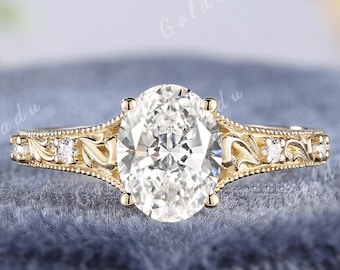 May include: A gold ring featuring a large oval-cut center stone and smaller round accent stones along the band. The band displays intricate engraved leaf-like patterns and milgrain detailing. The text GOLDENXANADU is visible at the top.