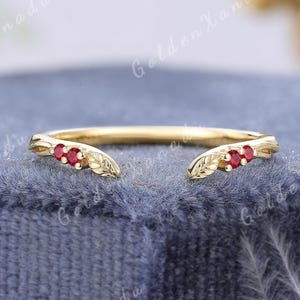 May include: A gold open-style ring featuring two small leaf-shaped accents on each end, each set with two round red gemstones. The band has a polished finish and the text GOLDENXANADU is visible in the background above the jewelry piece.