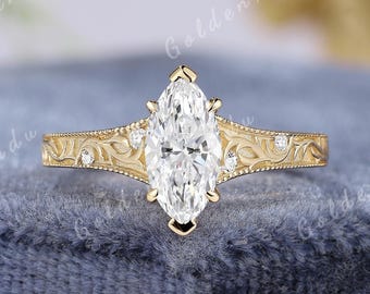 May include: A gold ring featuring a central marquise-cut clear gemstone held by six prongs. The band displays intricate engraved scrollwork patterns and is accented with four small round clear stones. The text GOLDENXANADU appears above the ring.