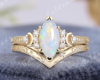 May include: A gold ring set featuring a marquise-cut opal center stone flanked by small round diamonds and crescent moon accents. The band includes a matching chevron-shaped wedding band with engraved star details and scrollwork patterns.