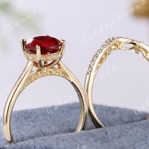 May include: A gold engagement ring set with a large red gemstone and a matching band with small diamonds. The rings are displayed in a blue velvet ring box.