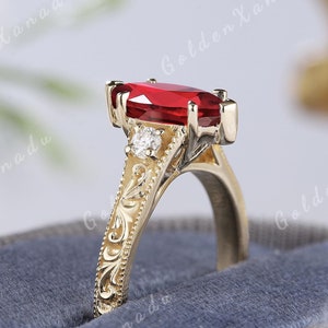 May include: A gold ring featuring a red oval gemstone and smaller diamond accents. The ring band is adorned with an intricate floral design.