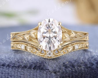Puede incluir: Un juego de anillos de compromiso de oro con un diamante de talla ovalada y una banda a juego. Los anillos presentan intrincados diseños florales y de hojas con pequeños diamantes de acento. El texto "GOLDENXANADU" está en la parte superior de la imagen.