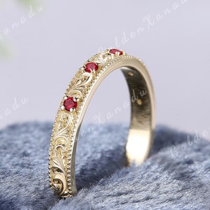 Könnte beinhalten: Ein goldener Bandring mit einem filigranen Blumendesign und fünf roten Edelsteinen. Der Ring liegt auf einem blauen Samtstoff.