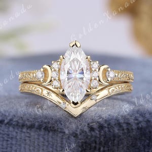 May include: A gold ring set featuring a marquise-cut center stone flanked by small round stones and crescent moon accents. The band includes engraved scrollwork and star details. The text GOLDENXANADU is visible at the top of the image.